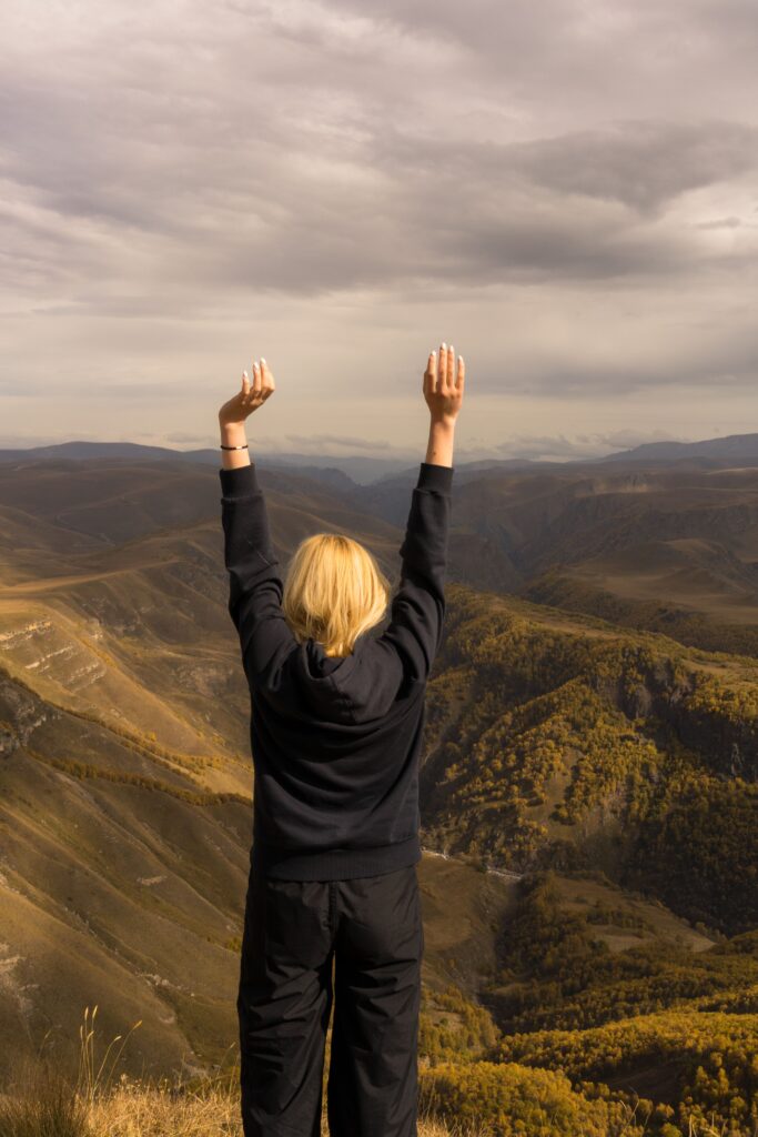 Back view of anonymous female standing with raised arms and admiring picturesque scenery of mountains in cloudy day
